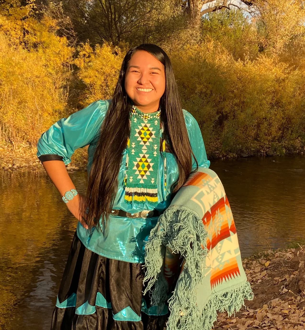 A smiling Cobell Scholar in a long beaded necklace and ribbon dress, a blanket over her arm, beside a river in autumn light.
