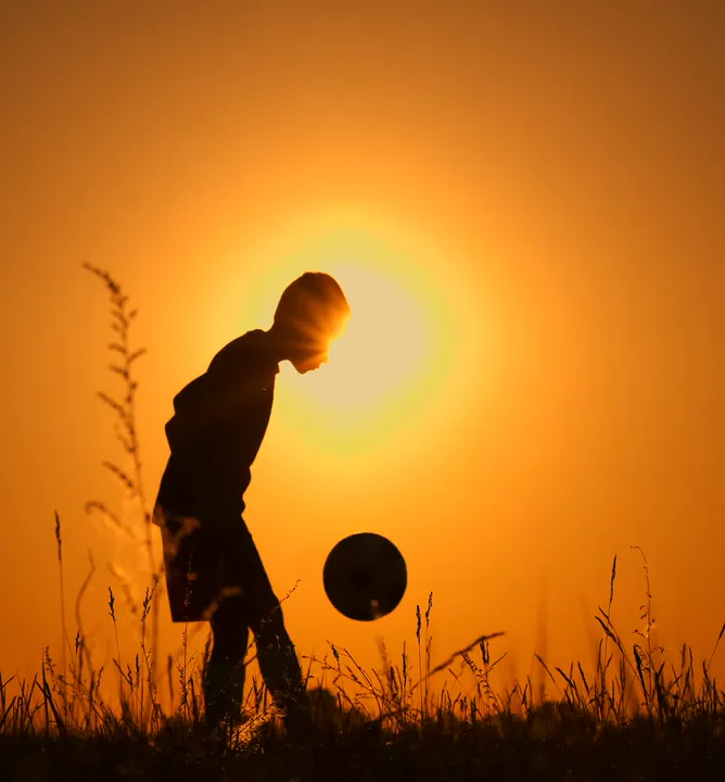 A boy juggles a football, silhouetted against the setting sun.