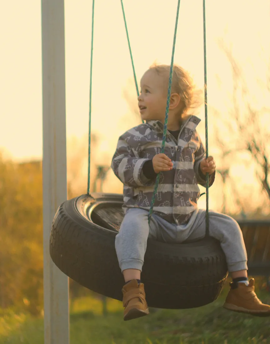 A smiling toddler rides a tyre swing in low golden sunlight.