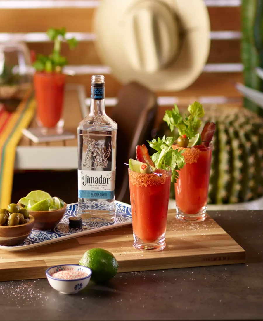 Bloody Marias with celery and limes beside a bottle of el Jimador on a sunny table.