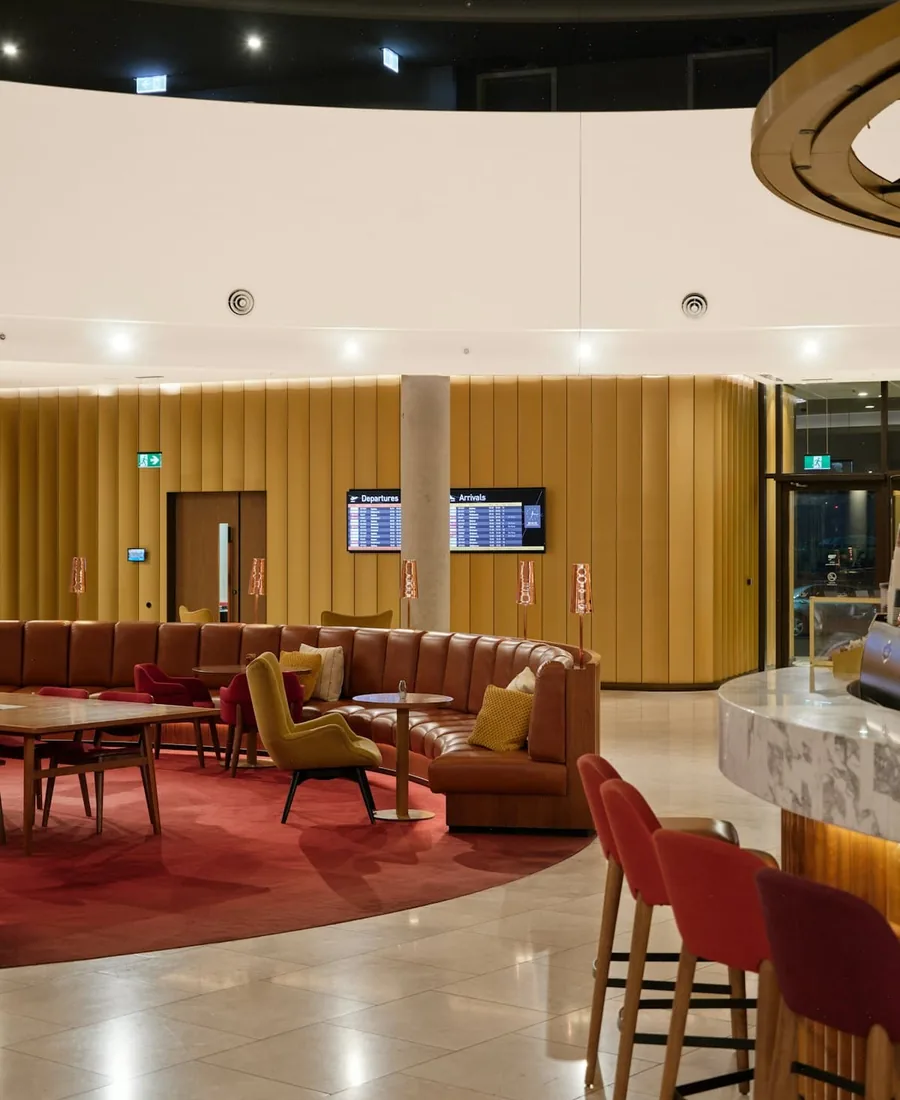 A terminal lounge with curved leather banquettes, a red carpet and a bar, departure screens on the wall.