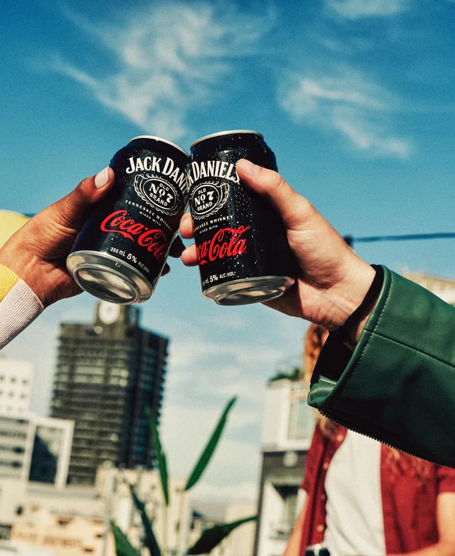 Two hands clink cans of Jack Daniel’s & Coca-Cola against a blue sky above a city rooftop.