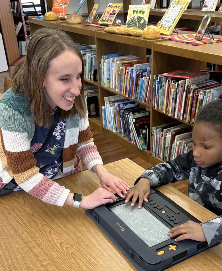 In a school library, a teacher and a child read together on a braille display.