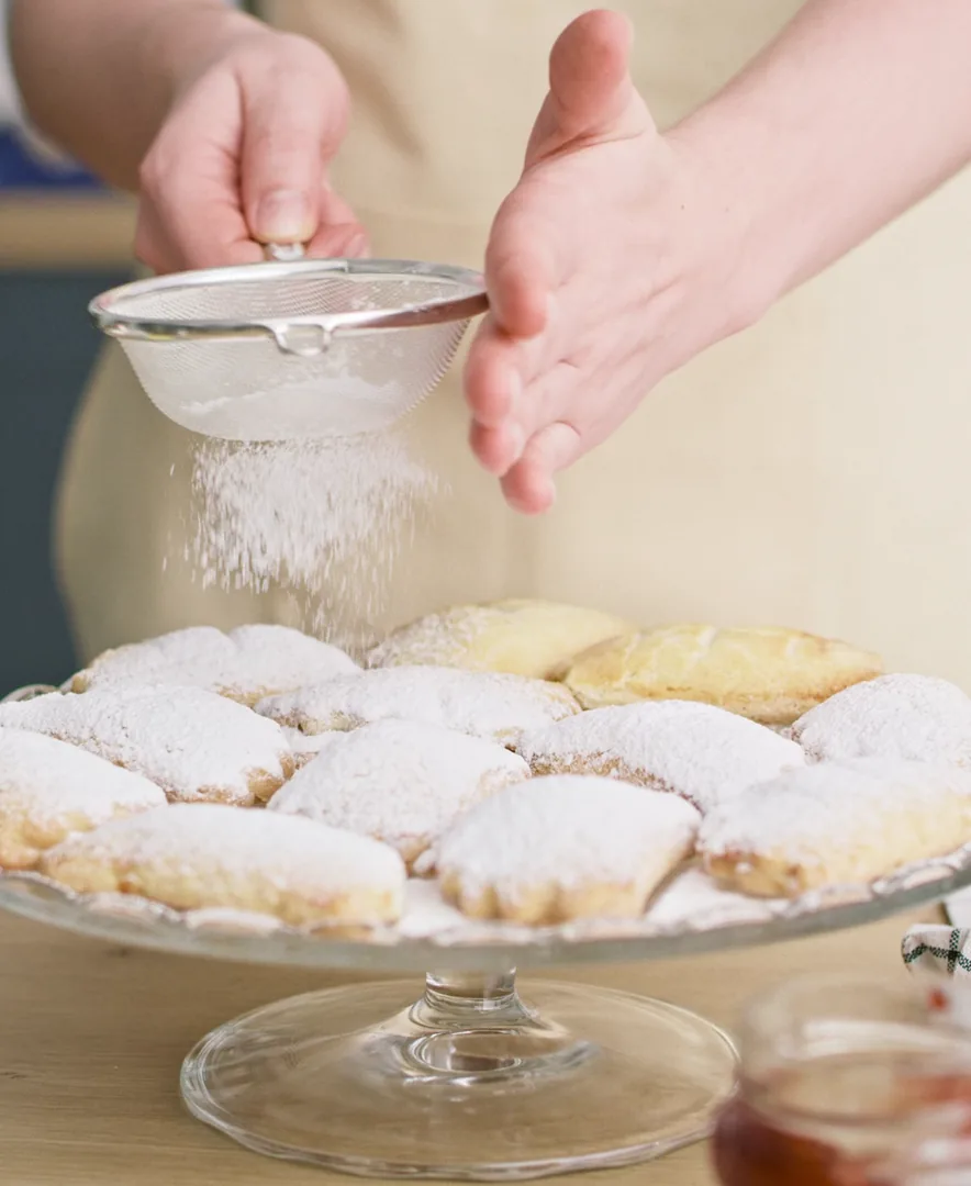 A baker sifts powdered sugar over pastries on a glass cake stand.