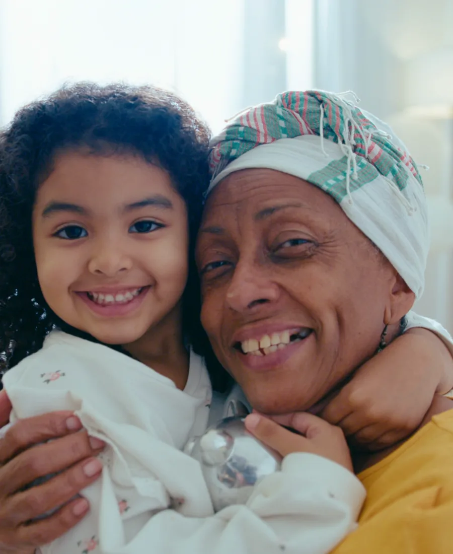 A smiling grandmother in a head wrap hugs her young granddaughter cheek to cheek on a sofa by a bright window.