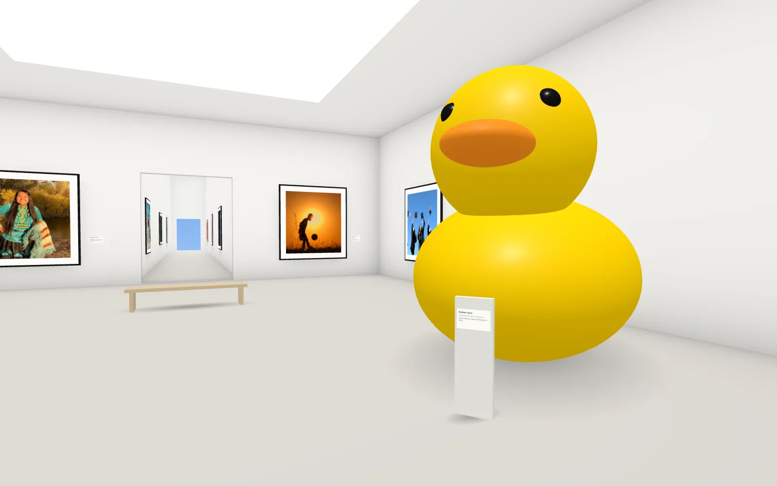 Inside the gallery: a white hall with framed work on the walls, a long corridor ending in a door open to the sky, and a giant yellow rubber duck