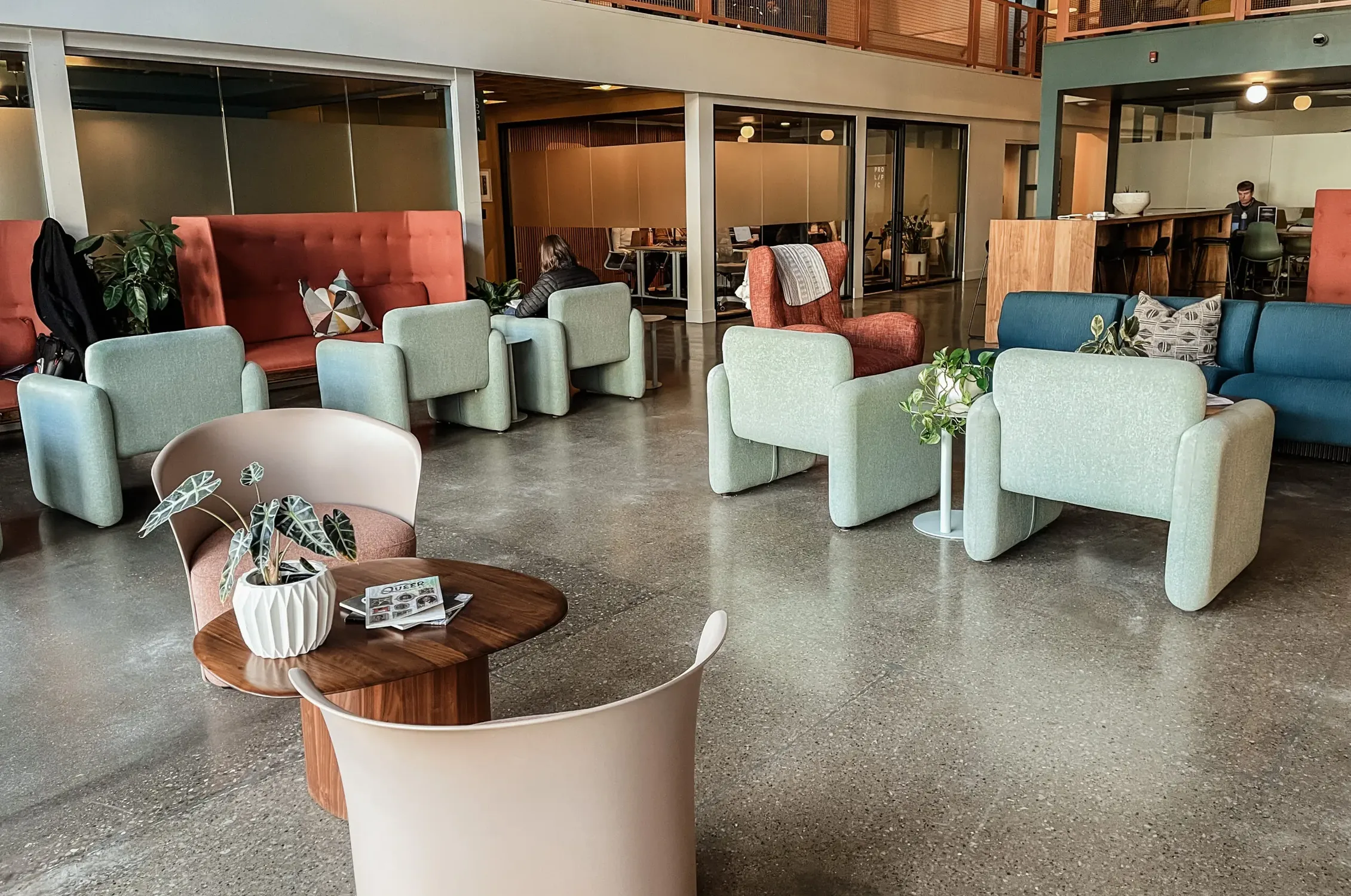 The ground-floor lounge: pastel armchairs, red sofas and a low wooden table, with glass-walled offices and the mezzanine above