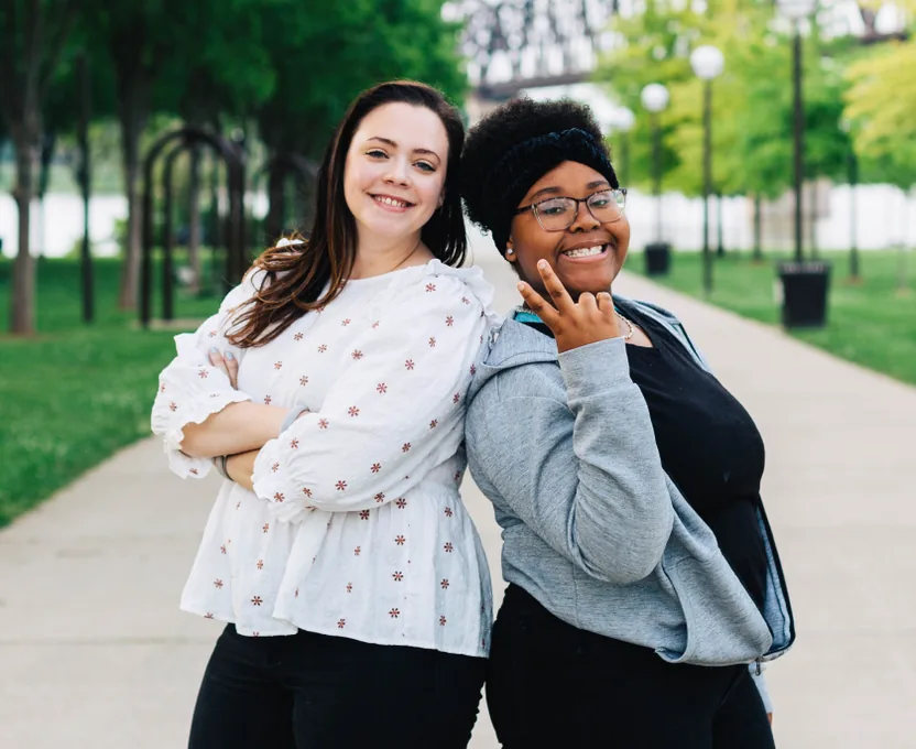 Two smiling women stand back to back on a tree-lined path.