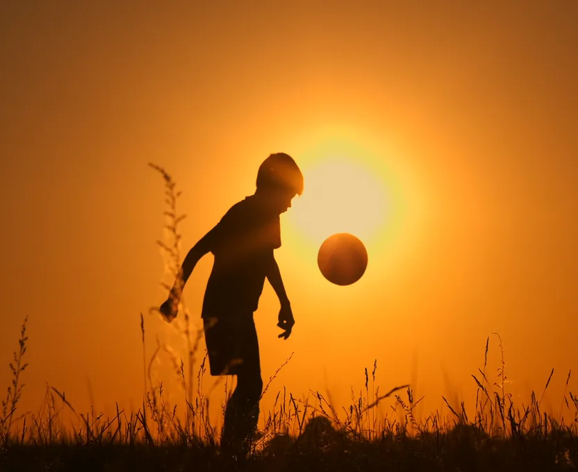 A boy juggles a football, silhouetted against the setting sun.