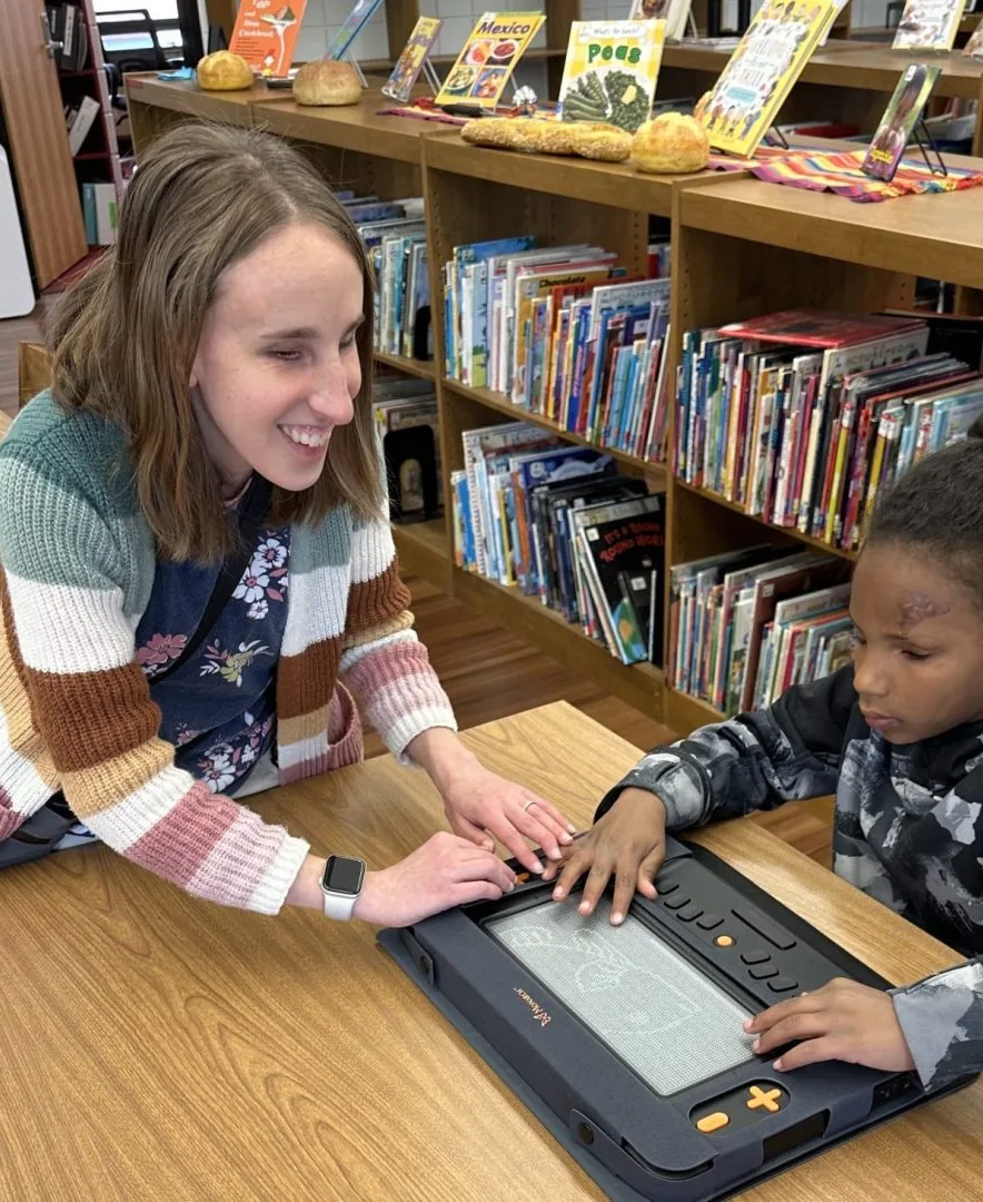 In a school library, a teacher and a child read together on a braille display.